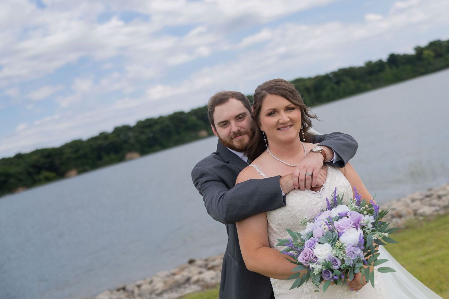 Bride and groom embrace near a lakeside railing, bride holding purple bouquet, groom in gray suit, on their wedding day.