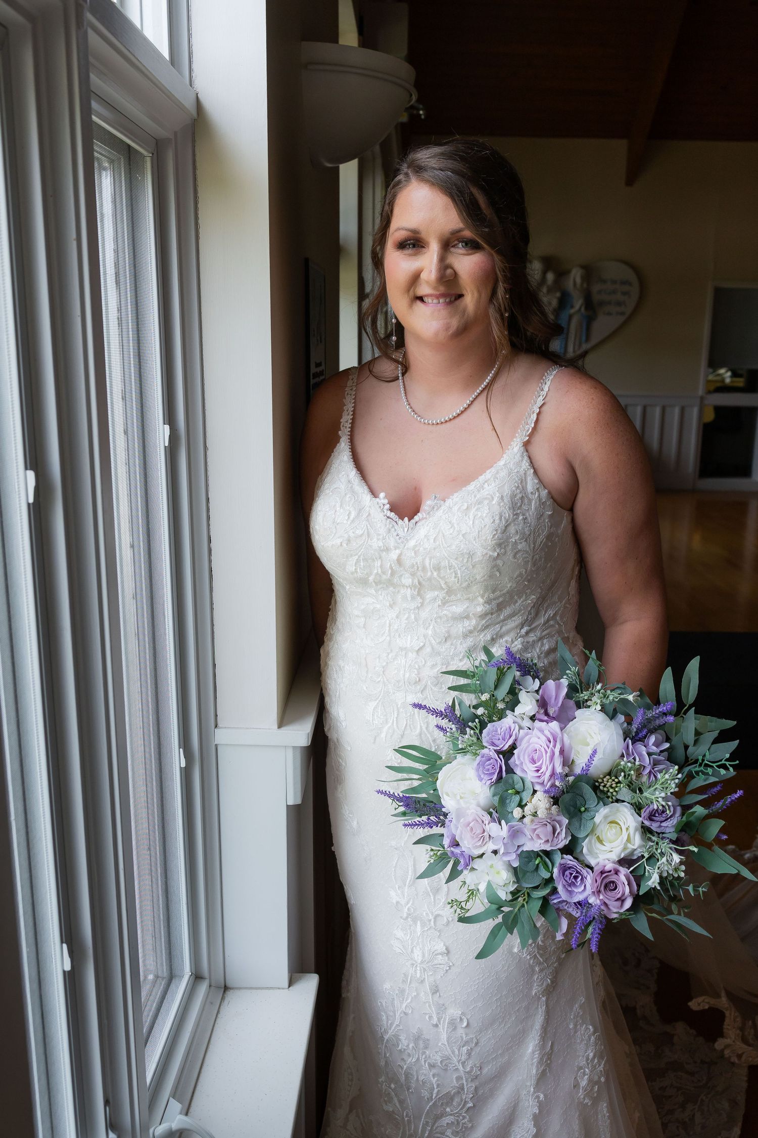 Bride in white lace gown holding purple and white bouquet, standing by window in elegant indoor setting.