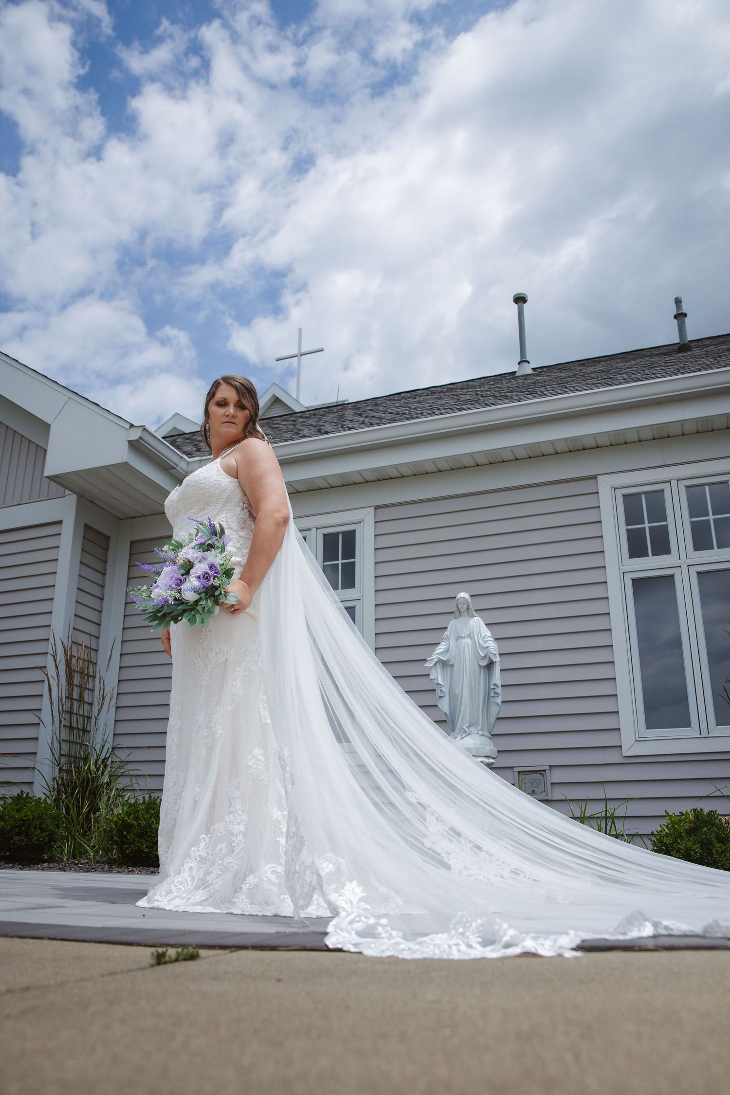 Bride in white lace gown with long veil stands outside a church with a floral bouquet under a cloudy sky.