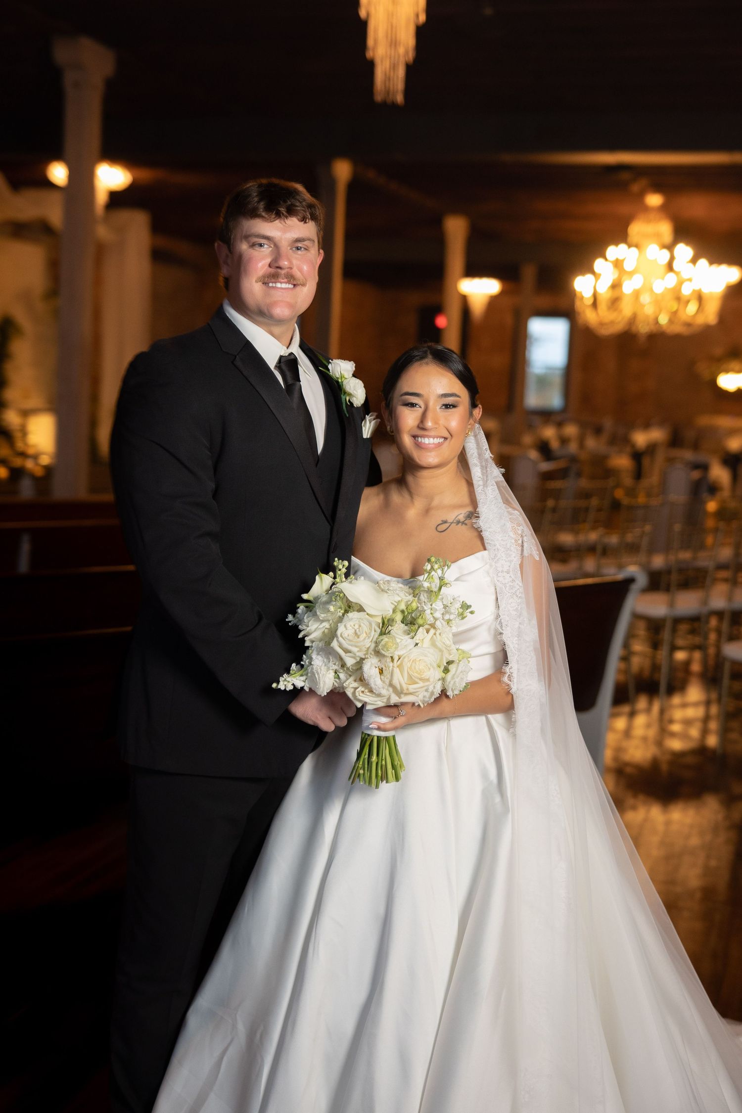 A newlywed couple poses with white bouquet in an elegant reception venue with chandeliers.