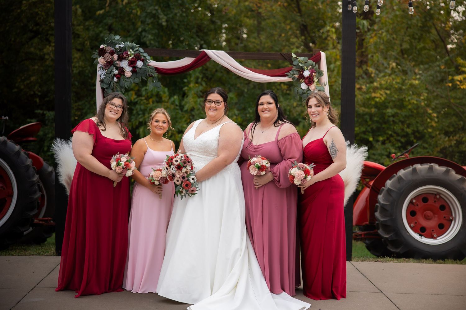 A wedding party poses outdoors wearing burgundy and pink dresses against a natural backdrop.