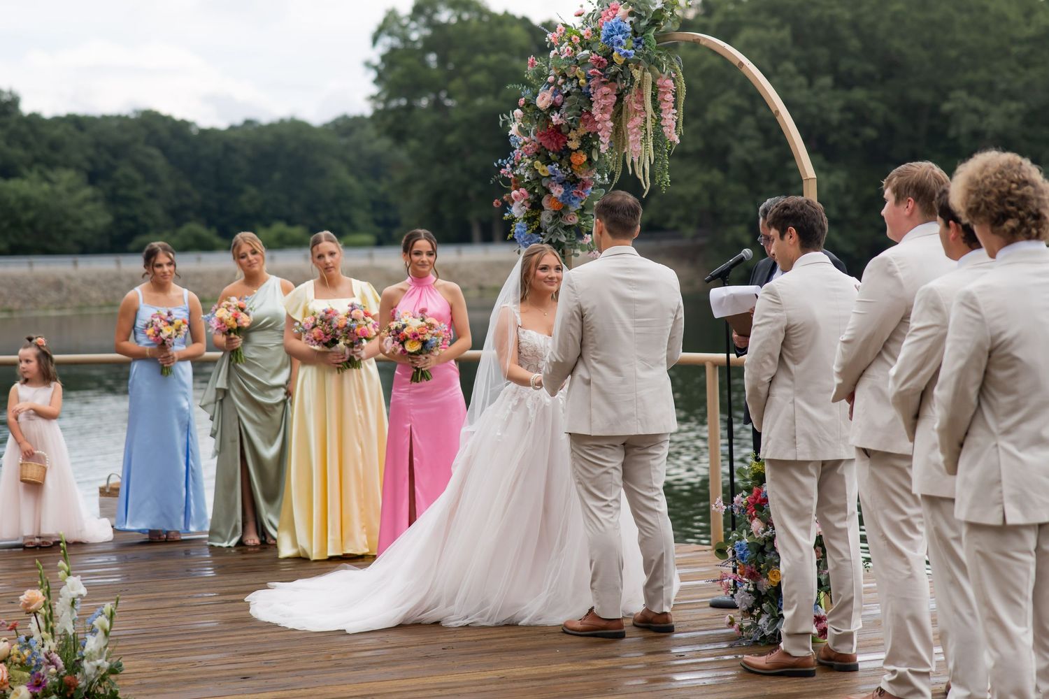 Outdoor wedding ceremony on a dock with bride, groom, officiant, and colorful bridesmaids under a floral arch.