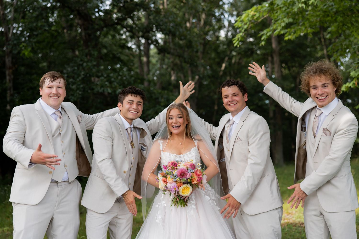 Bride in white gown poses with four groomsmen in gray suits outdoors at a wedding celebration.