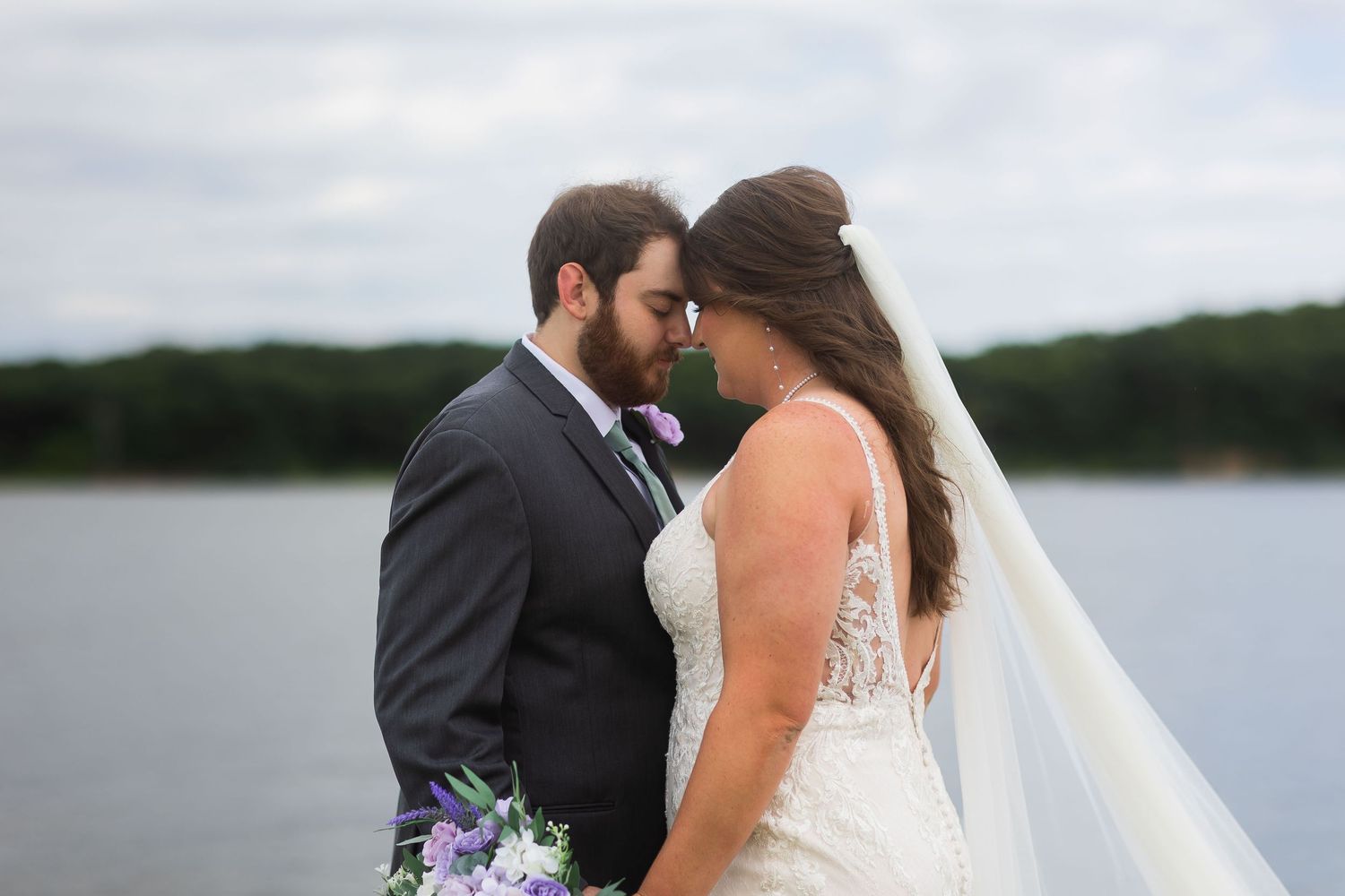 Bride and groom share a romantic moment by a lake, bride holding a purple bouquet.