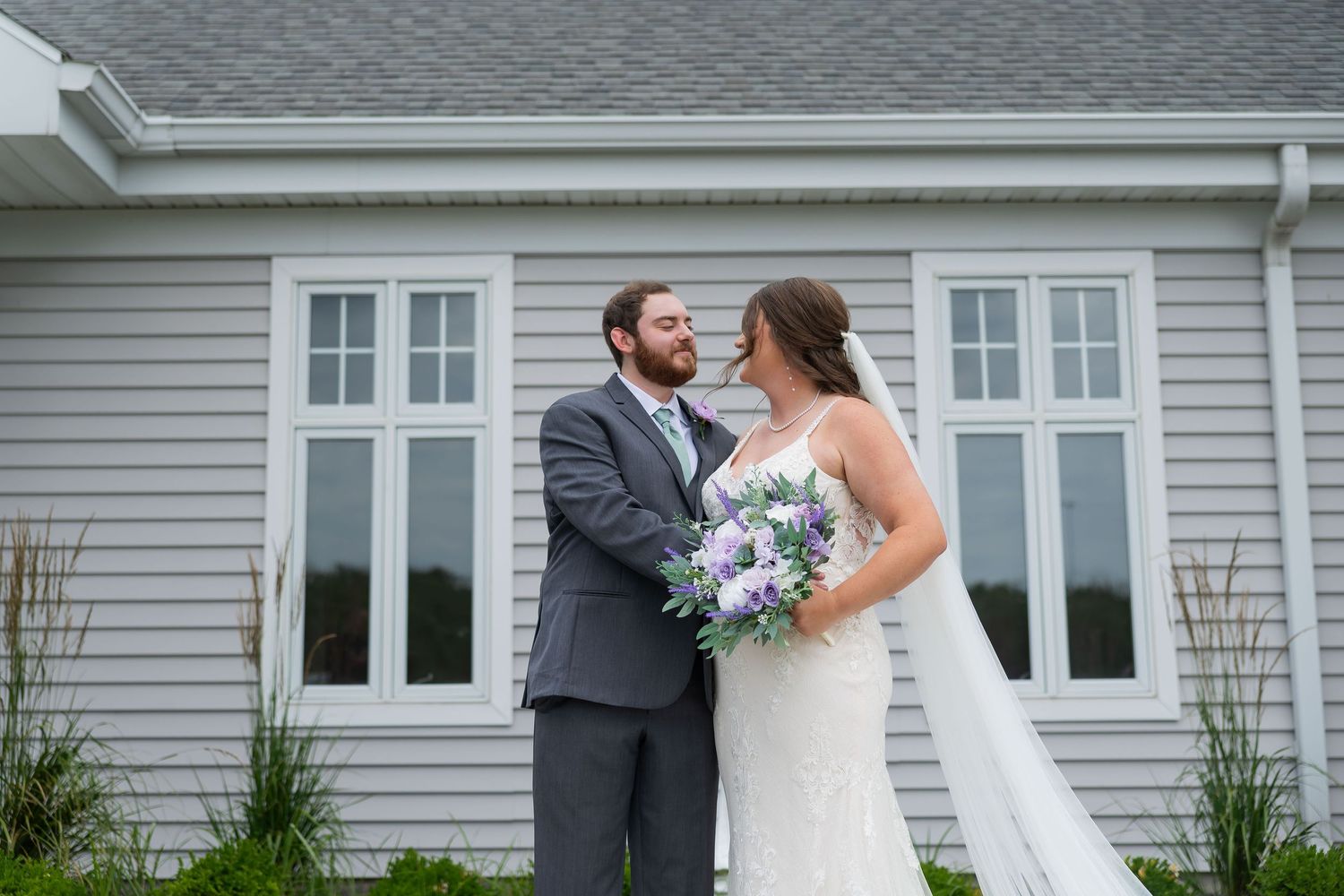 Smiling bride and groom sharing romantic moment outdoors holding purple floral bouquet at venue.