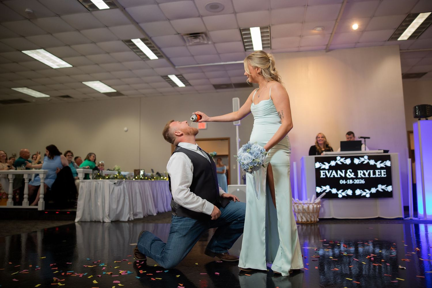 Groom kneels before bride on wedding dance floor with DJ booth displaying Evan and Rylee sign.