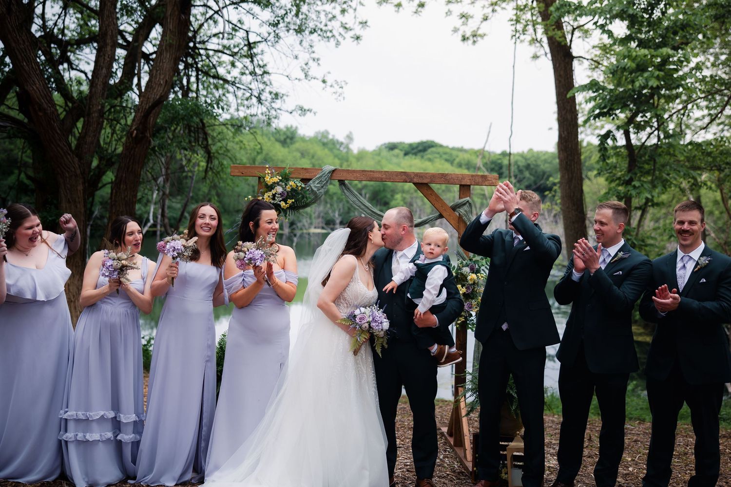 Joyful wedding party celebrates outdoors near water under a wooden arch with bridesmaids in lavender dresses.