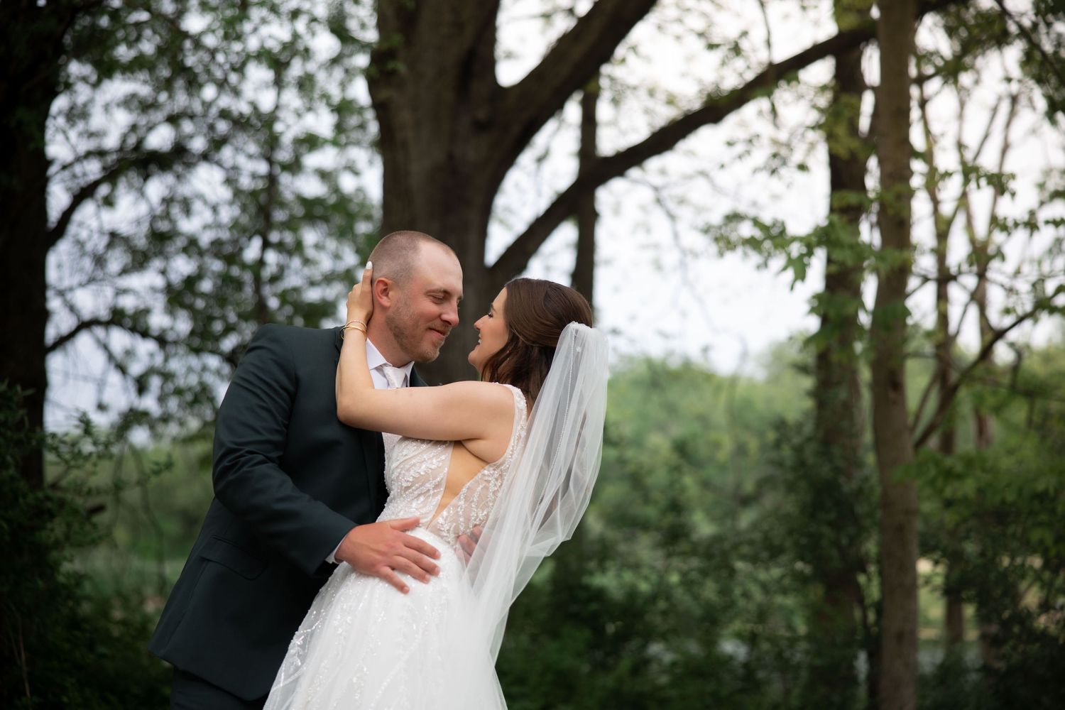 Bride and groom share a tender kiss surrounded by lush green trees at outdoor wedding.