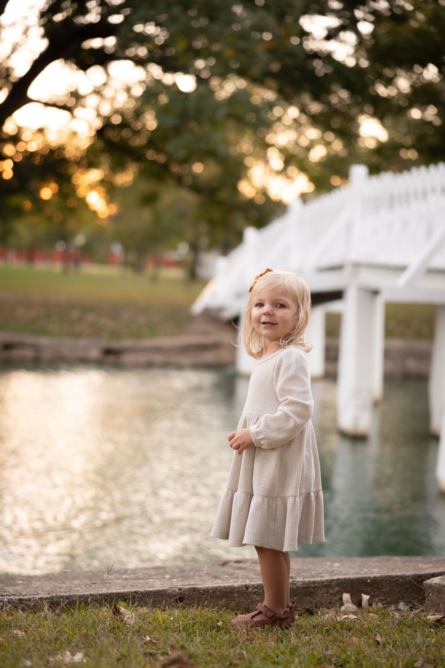 A small child in a cream dress stands by a pond with a white bridge in the background at sunset.
