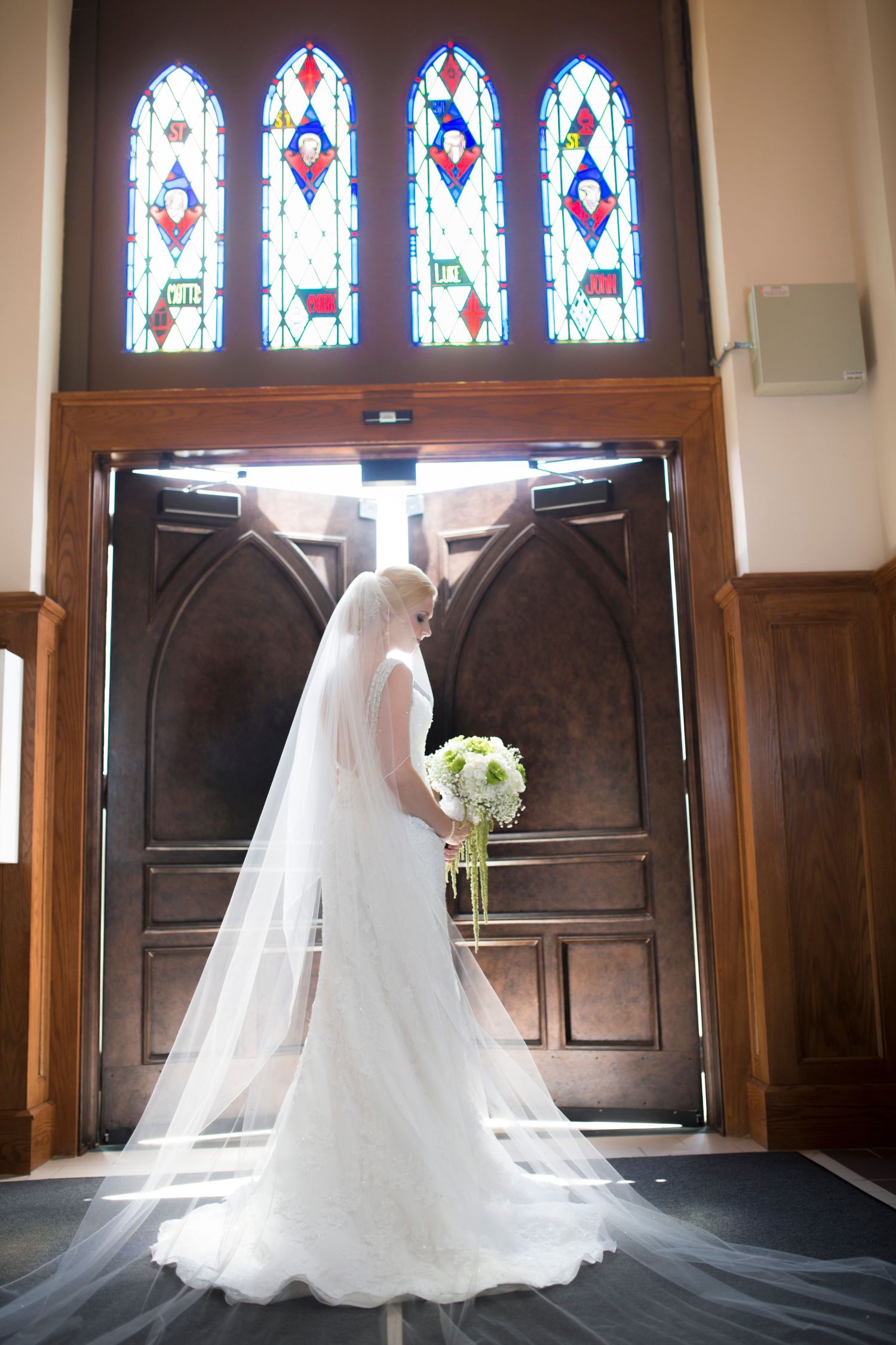 A bride in a flowing white wedding gown stands in a church with stained glass windows creating a dramatic silhouette.