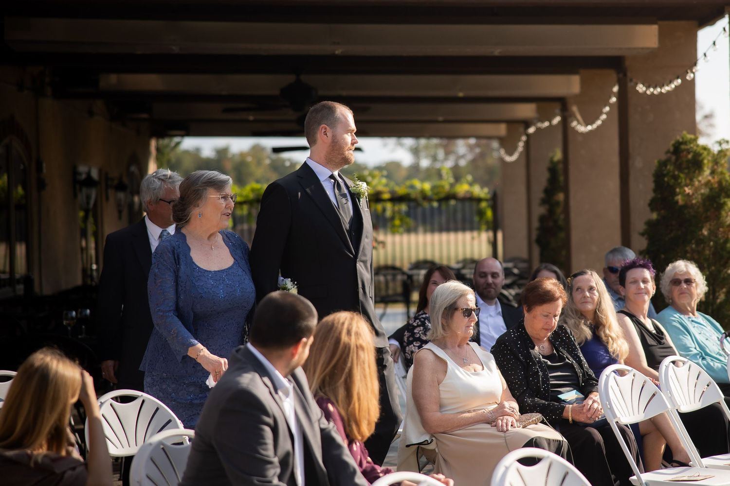 Wedding guests gather under a covered outdoor venue during a ceremony on a sunny day.