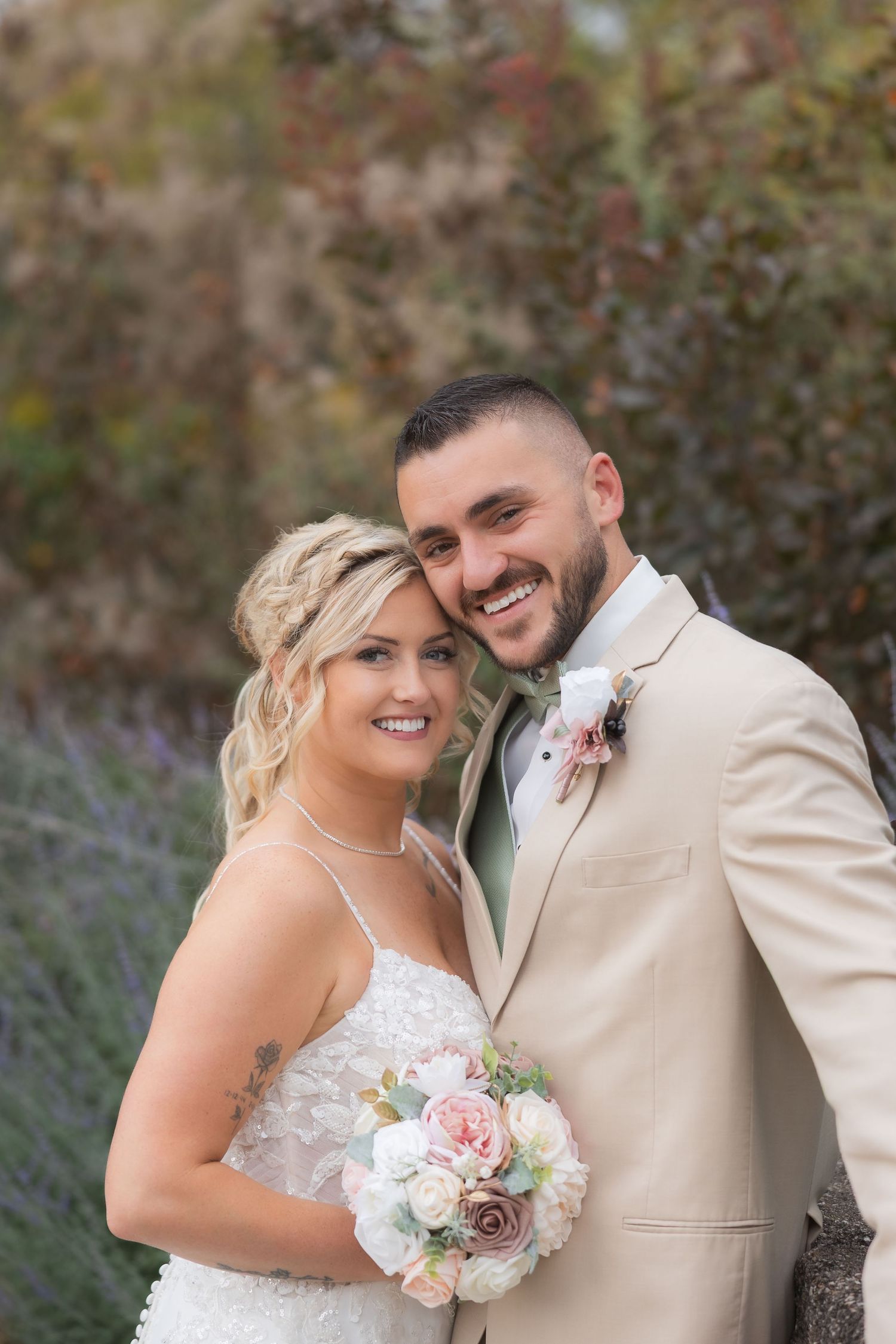 Couple in wedding attire posing with a pink and white floral bouquet against a garden backdrop.