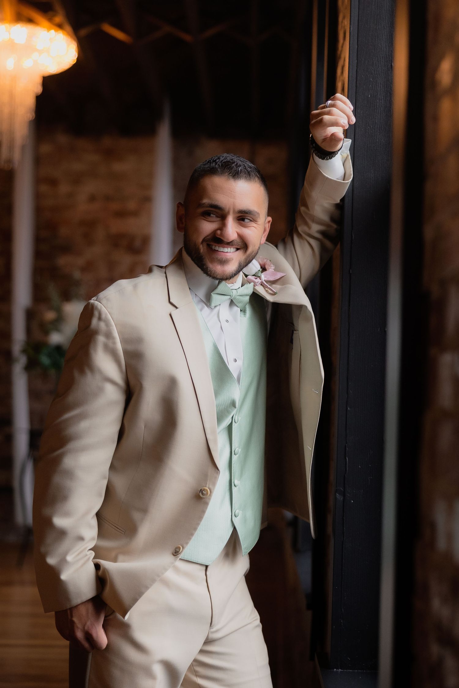 A groom in a tan suit with mint green vest and bowtie poses confidently against a rustic indoor backdrop.