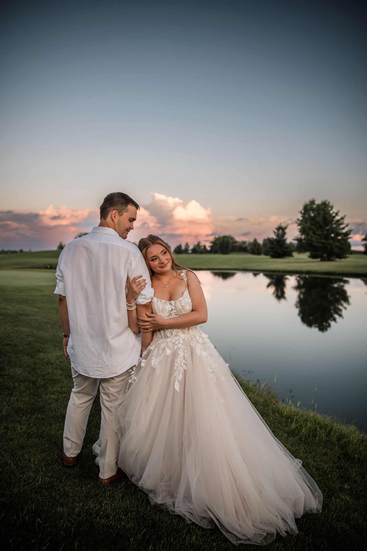 A bride and groom share a romantic moment by a reflective pond at sunset, with pink clouds and lush greenery.