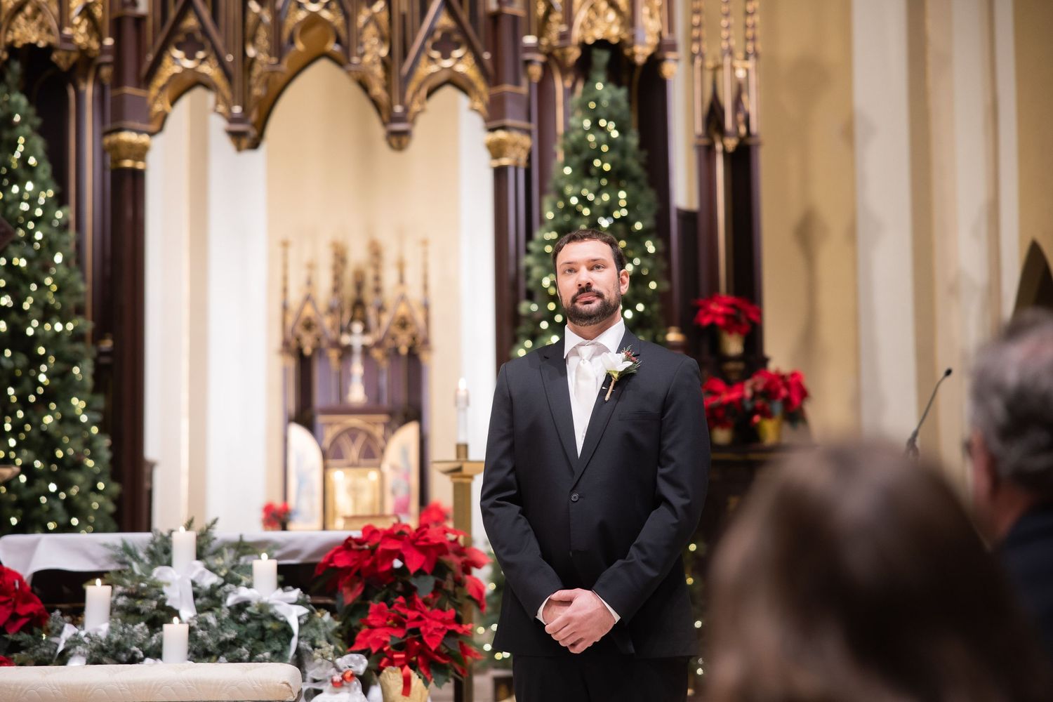 Groom in black suit standing at church altar decorated with Christmas poinsettias and trees.