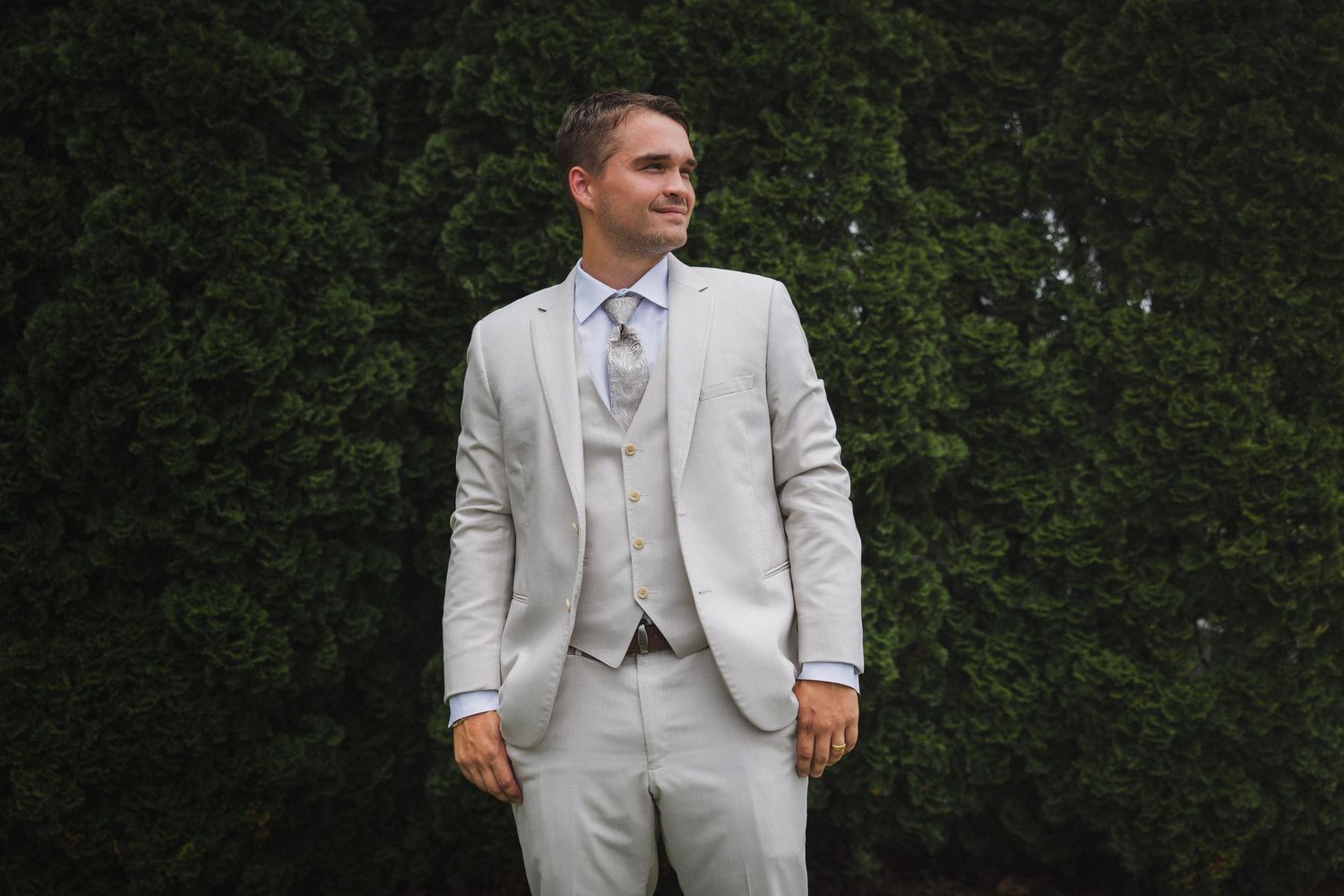 Handsome groom in light gray three-piece suit and blue tie posing outdoors in front of lush green hedges.