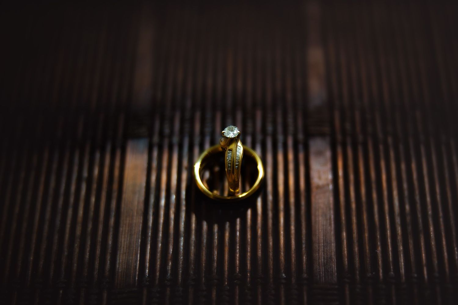 Two gold wedding rings with a diamond solitaire stacked together on a dark wooden surface.