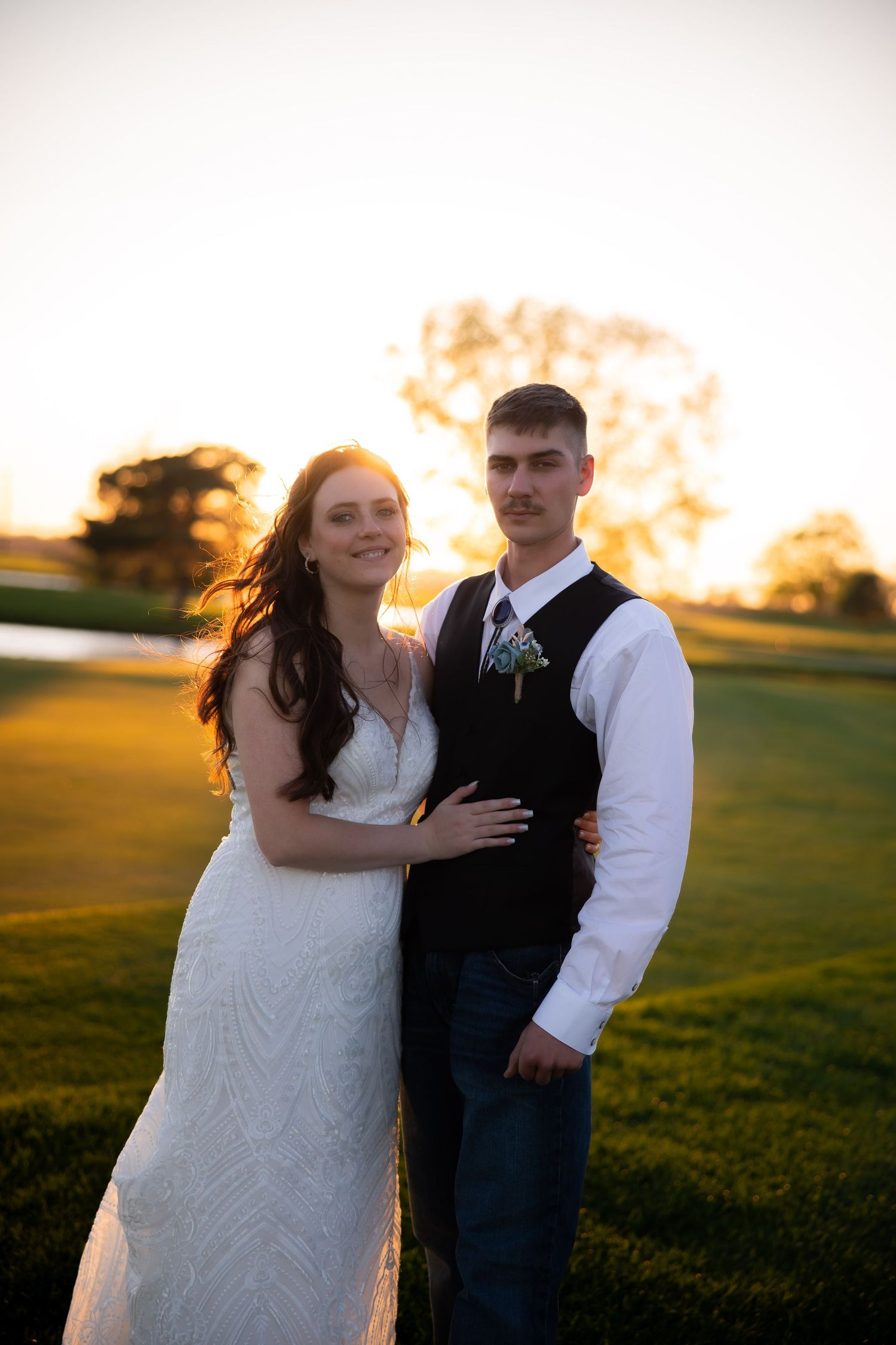 Bride in white lace gown and groom in vest embrace during golden sunset on wedding day.