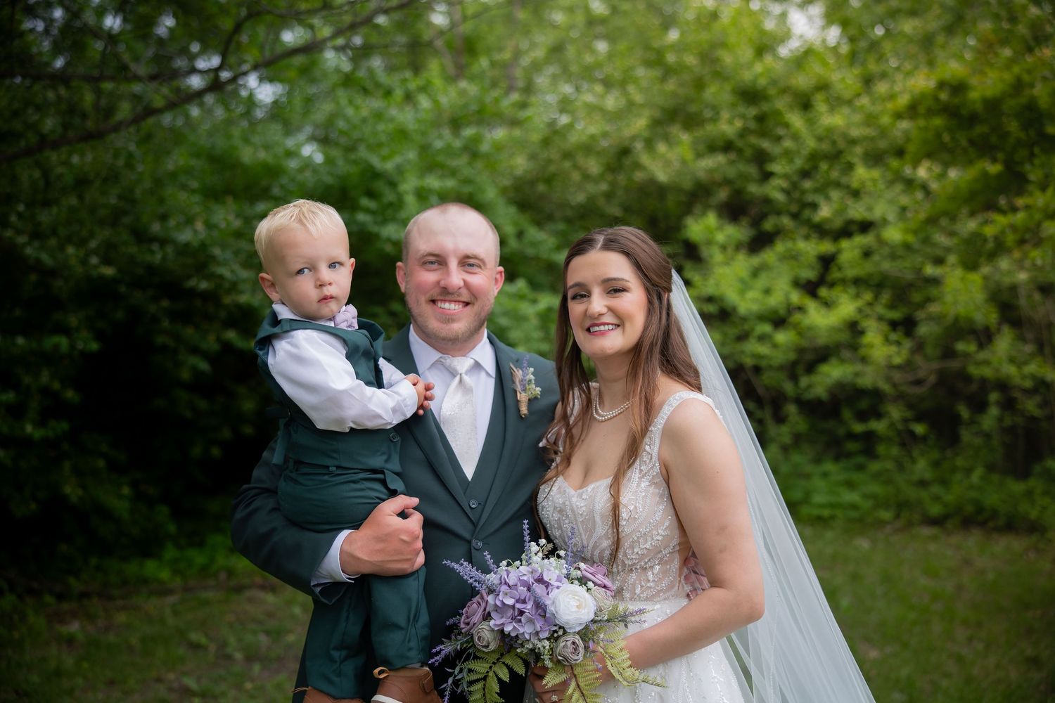 Groom in dark green suit holds toddler boy beside bride in white gown with veil holding purple bouquet outdoors.