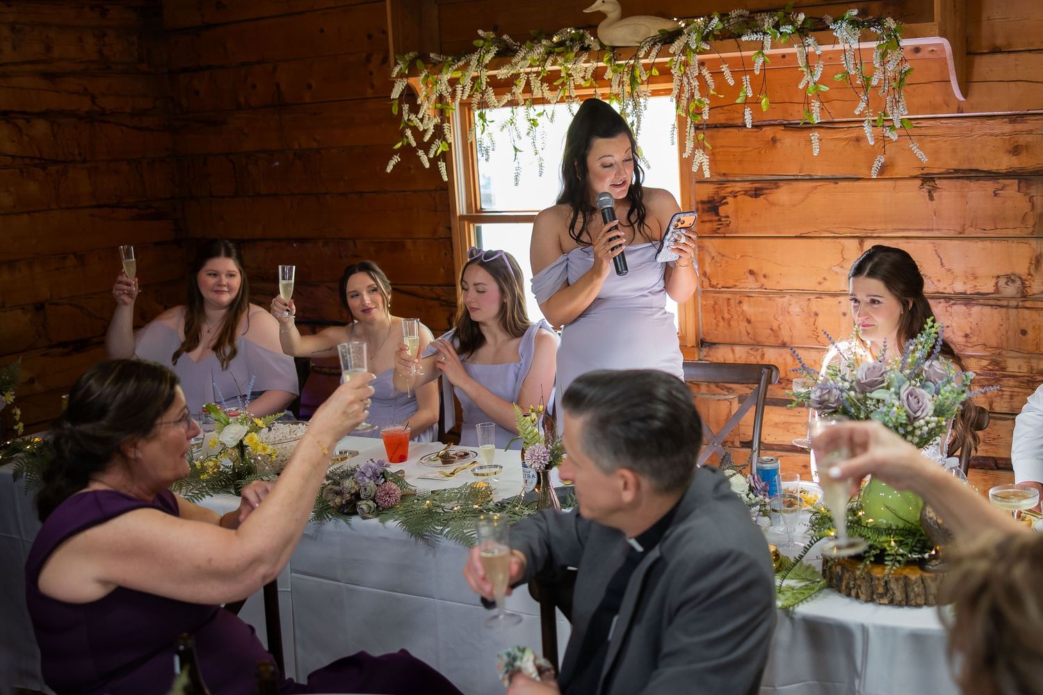Bridesmaid gives champagne toast speech at rustic barn wedding reception with bridal party raising glasses.