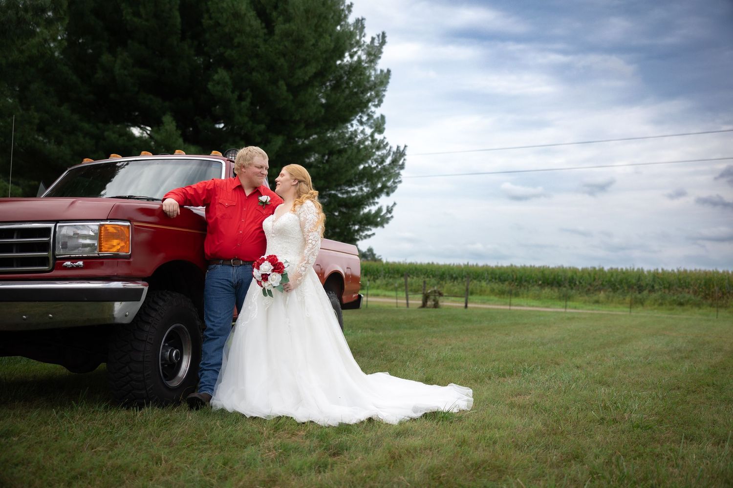 Bride and groom pose beside a red pickup truck in a green field on their rustic outdoor wedding day.