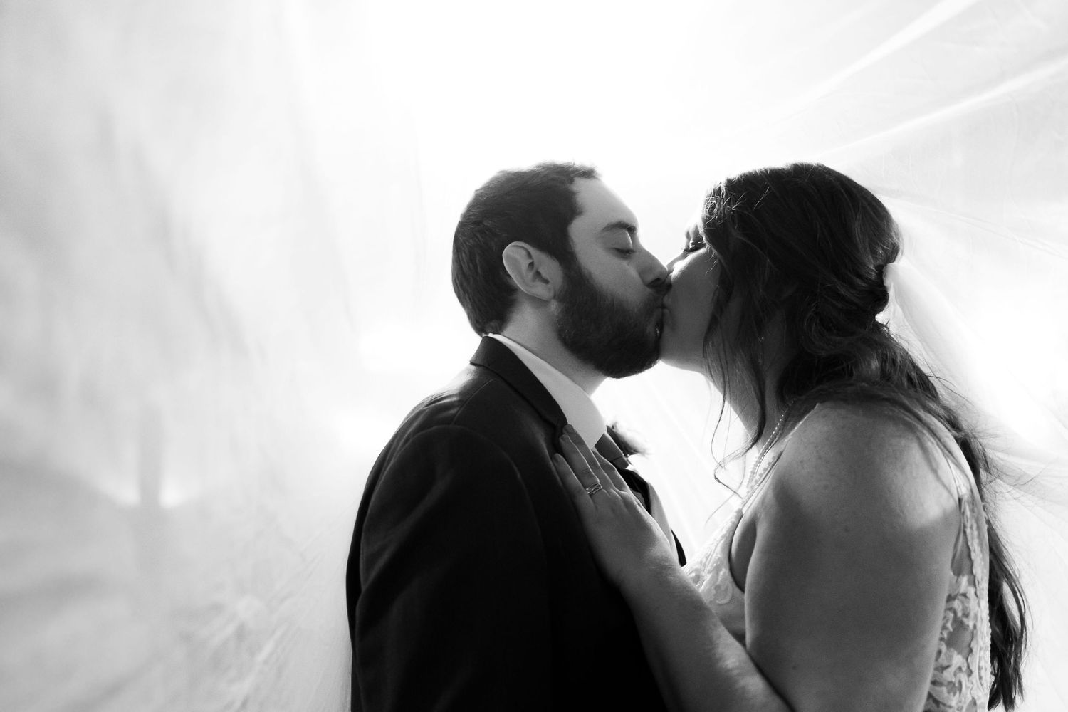 Black and white photo of a bride and groom sharing a romantic kiss beneath a flowing veil on their wedding day.