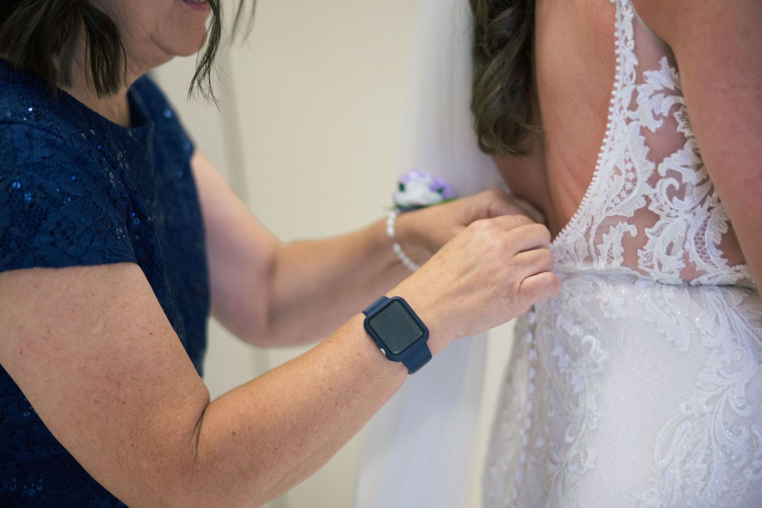 Woman in blue dress fastening buttons on a white lace wedding gown, wearing an Apple Watch.