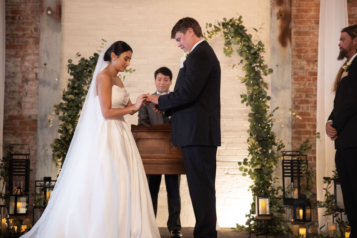 A wedding ceremony takes place in front of a brick wall adorned with greenery and lanterns.