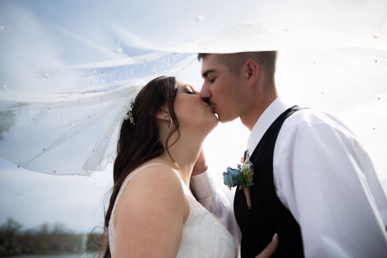 A bride and groom share a romantic kiss under a flowing white veil on their wedding day outdoors.