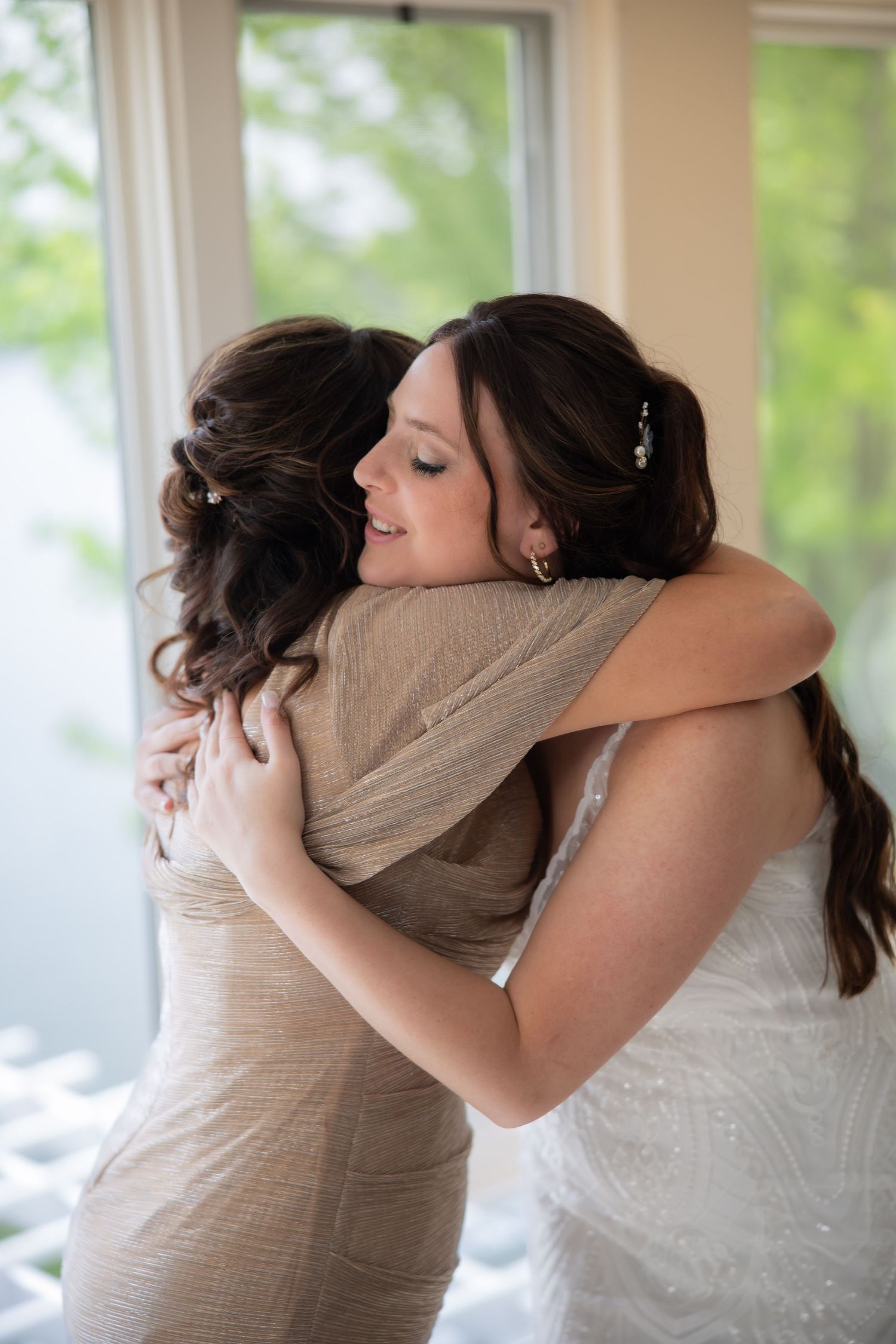 A bride in a white gown shares a heartfelt hug with a woman in a gold dress near a bright window.