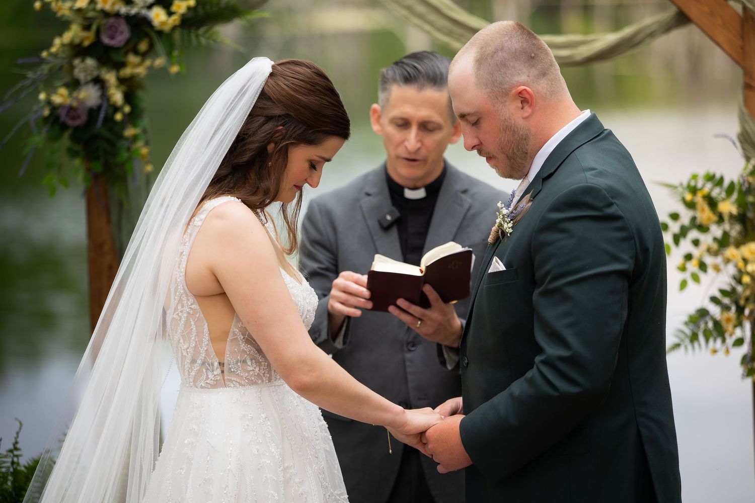 Bride and groom hold hands during vows with officiant reading from Bible at outdoor woodland wedding ceremony.