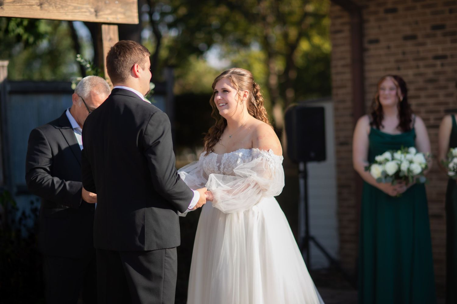 Bride and groom holding hands during outdoor wedding ceremony with bridesmaid in green dress in background.