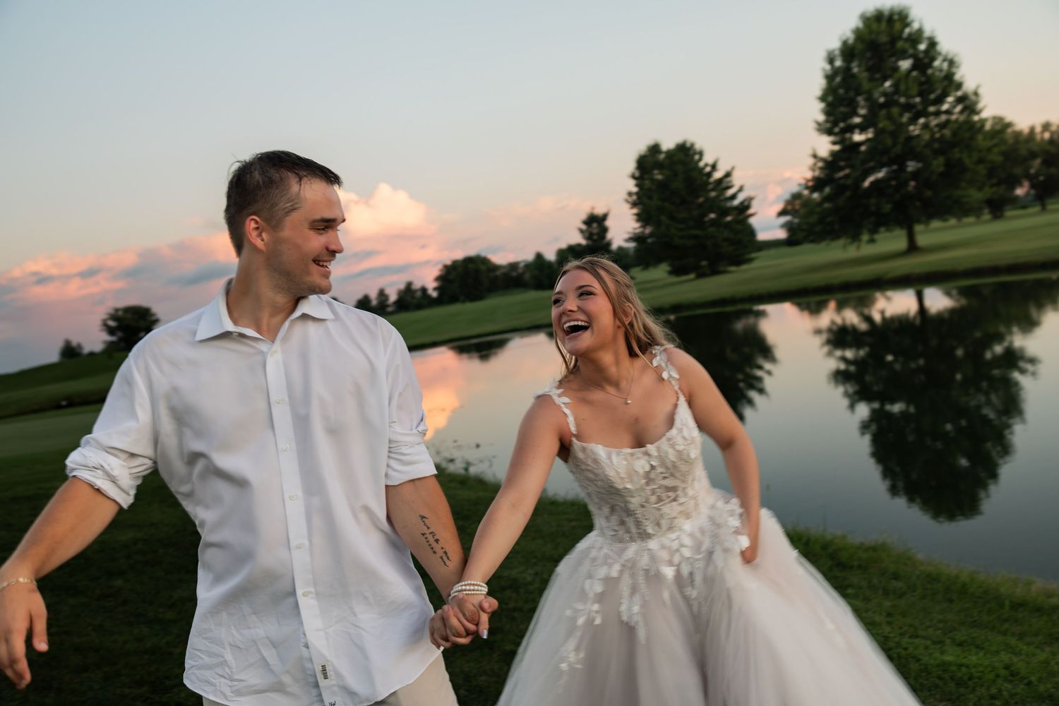 Bride and groom laugh while holding hands near a pond at sunset during their wedding reception.