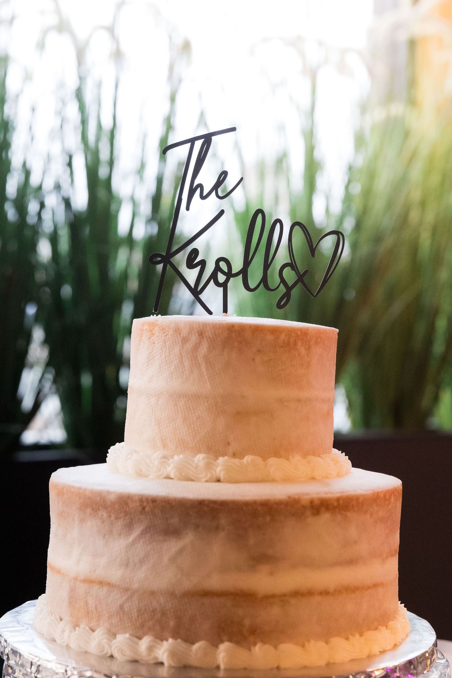 Three-tiered white wedding cake with elegant script topper displayed against natural greenery.