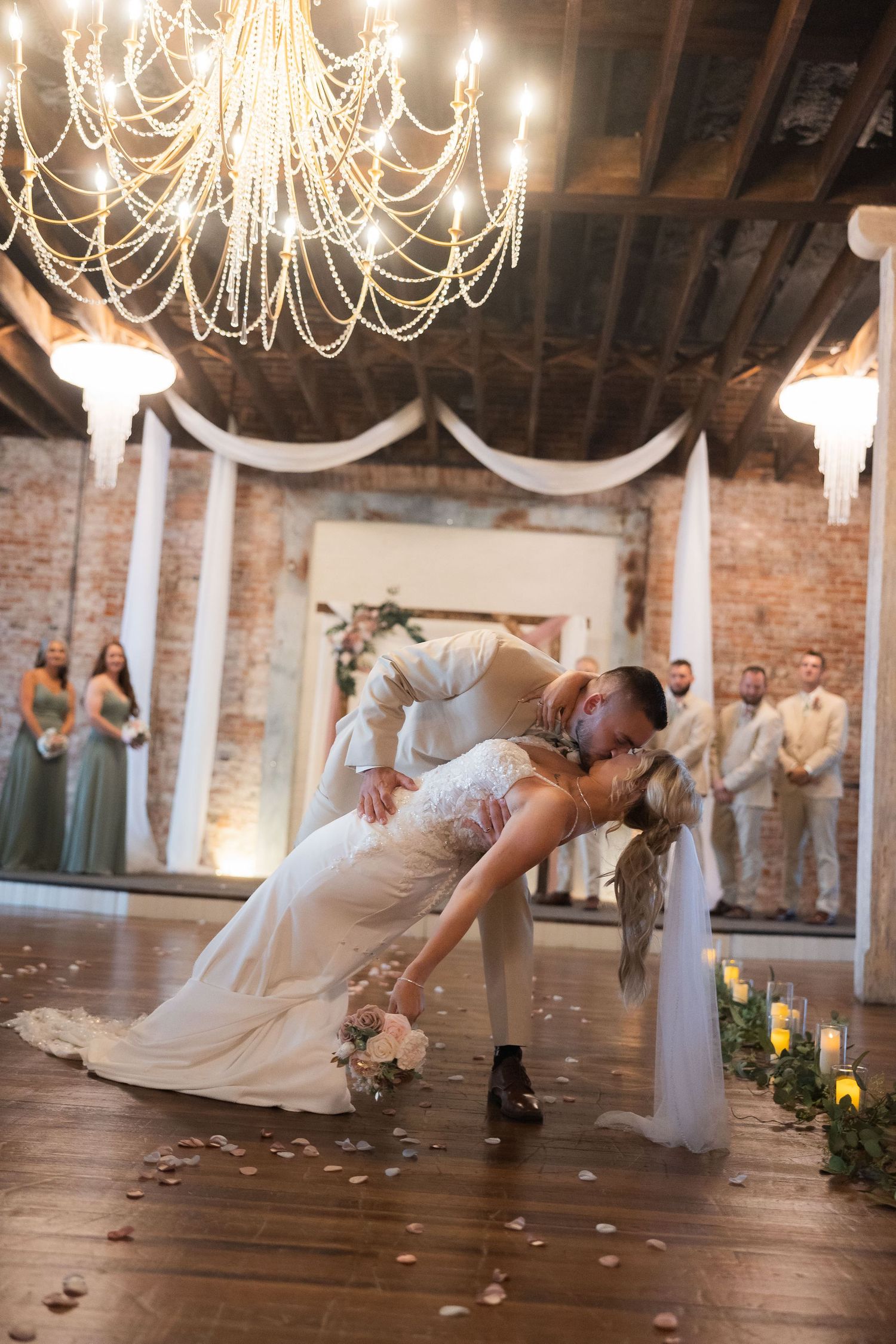 A wedding couple performs a romantic dip dance move under crystal chandeliers in a rustic venue.