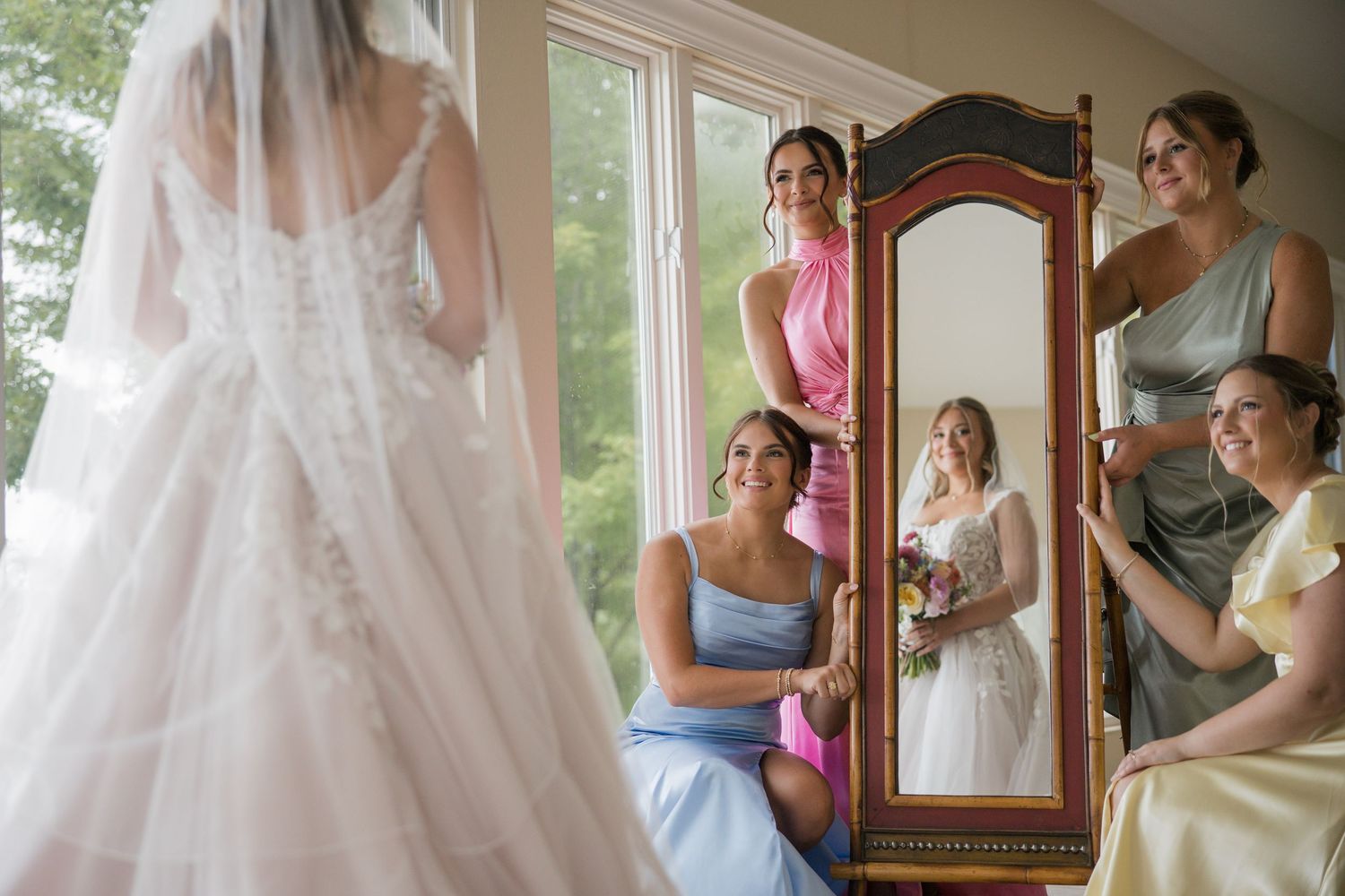 Bridesmaids in blue and pink dresses admiring bride in lace wedding gown reflected in ornate vintage mirror.