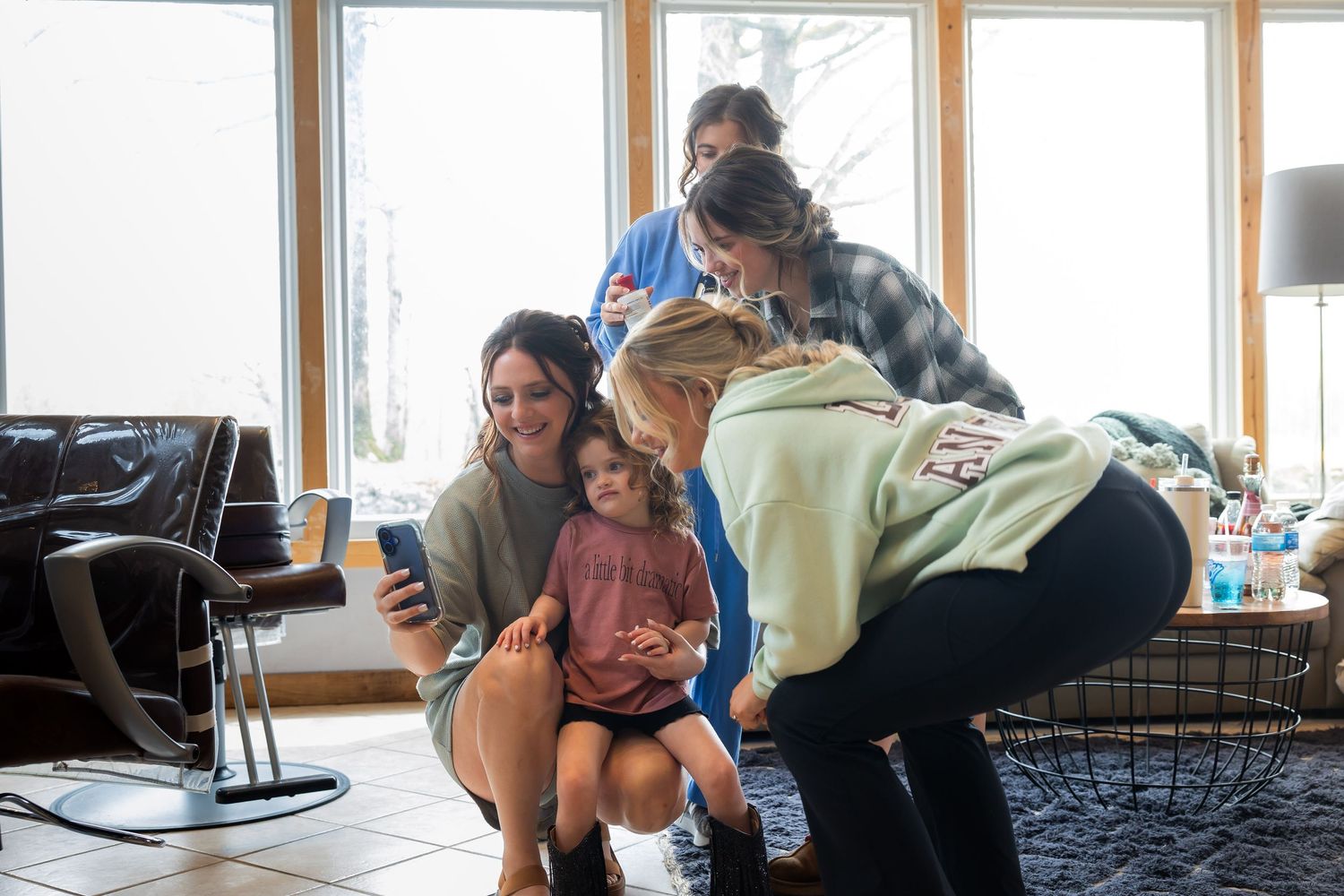 Group of women and children gathered indoors sharing a smartphone, smiling together in a bright living room.