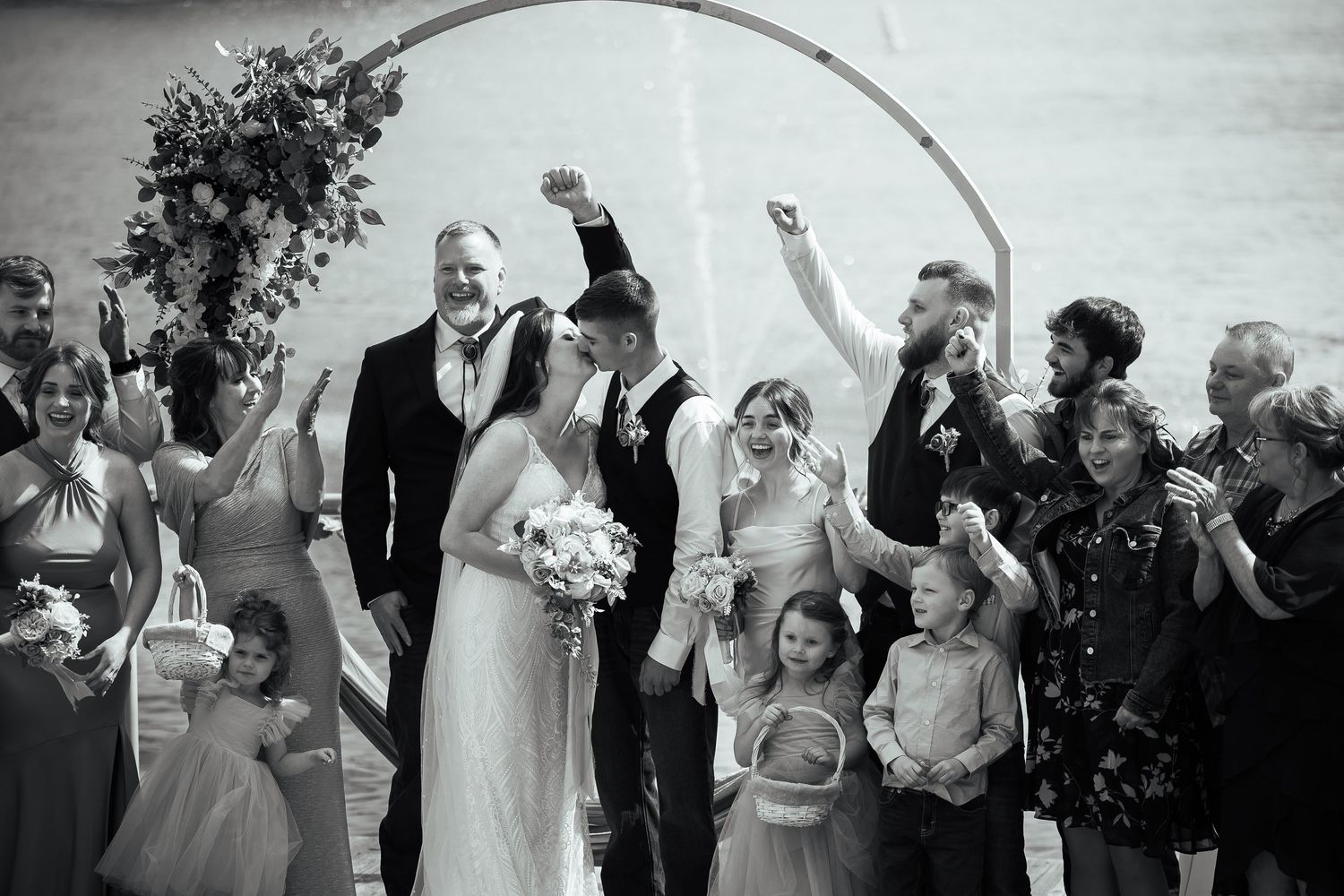 Black and white photo of joyful wedding party celebrating after outdoor ceremony under circular floral arch.