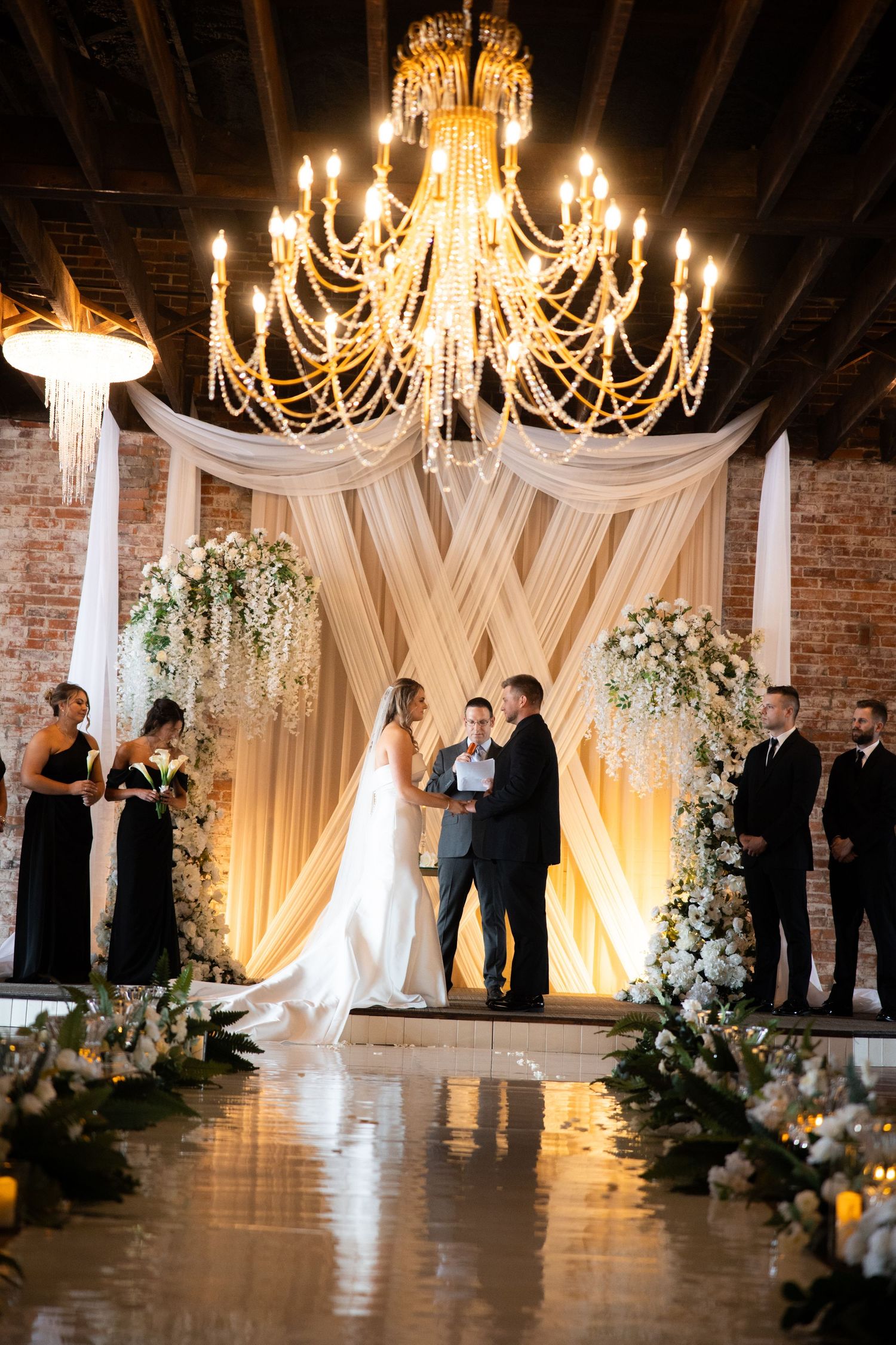 Romantic wedding ceremony venue with draped fabric, chandeliers, and white floral arrangements.