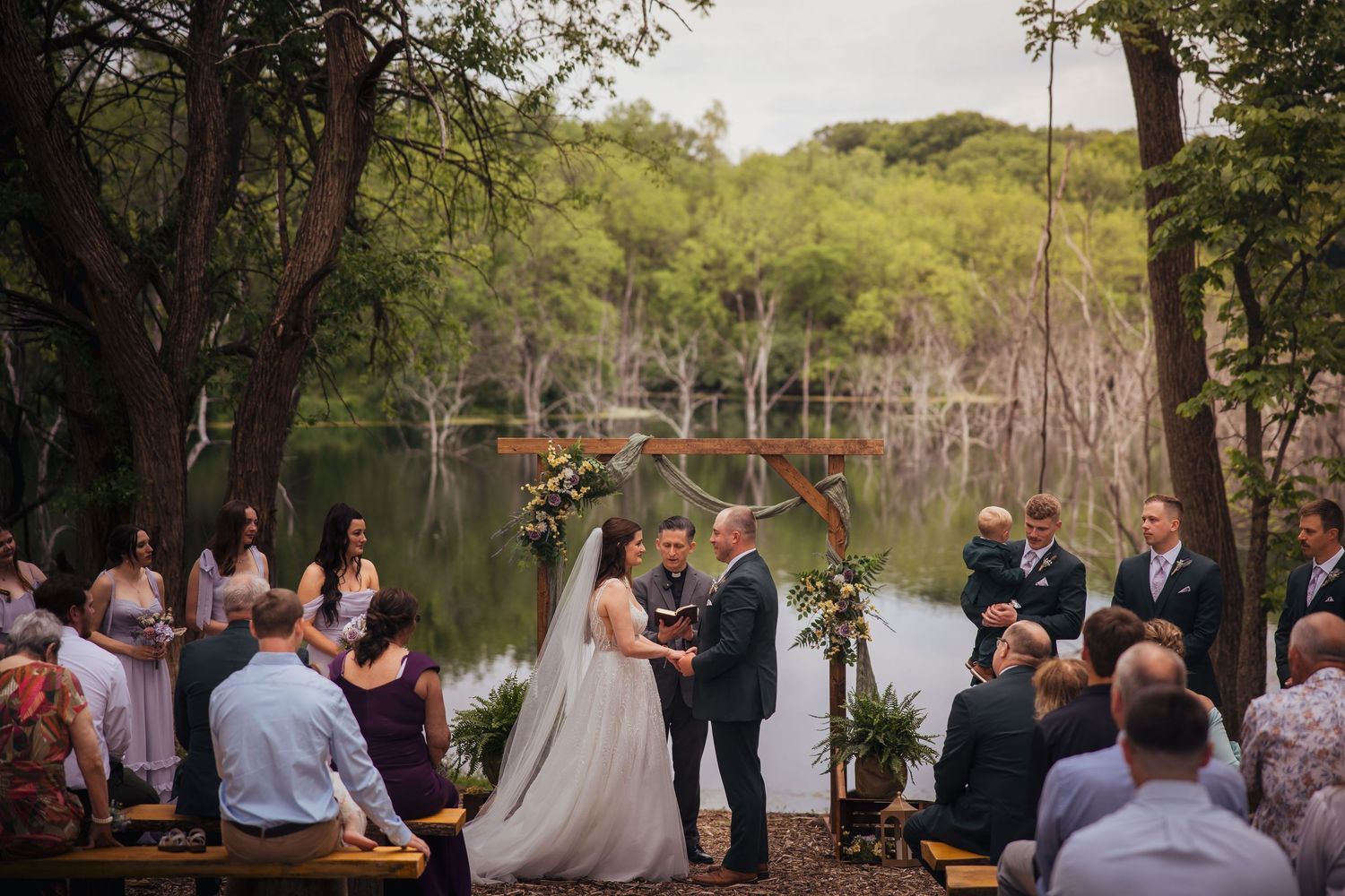 Outdoor wedding ceremony by a lake with guests seated and couple at floral arch.