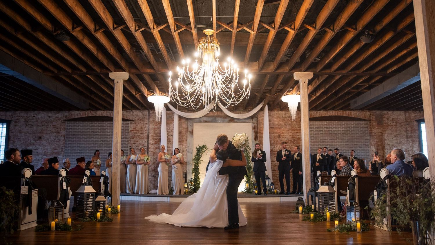 A couple shares their first dance under a crystal chandelier in a rustic venue with exposed wooden beams and candlelit aisle.