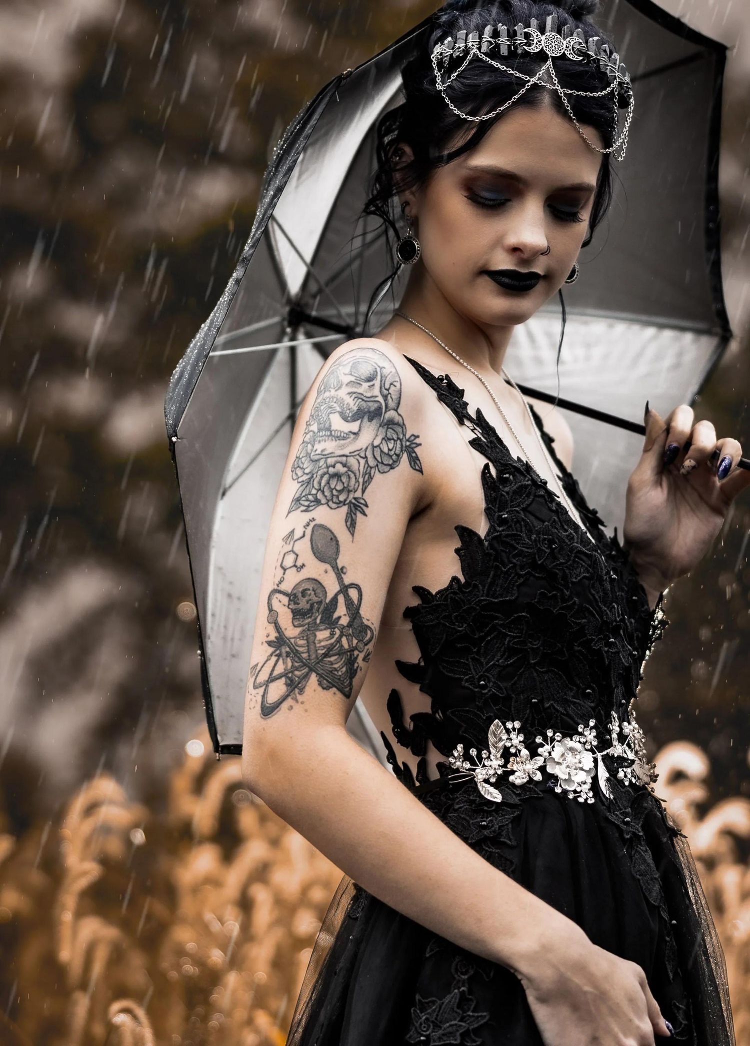 Gothic woman in black lace gown with floral arm tattoo holds umbrella in rain, wearing ornate silver headpiece.
