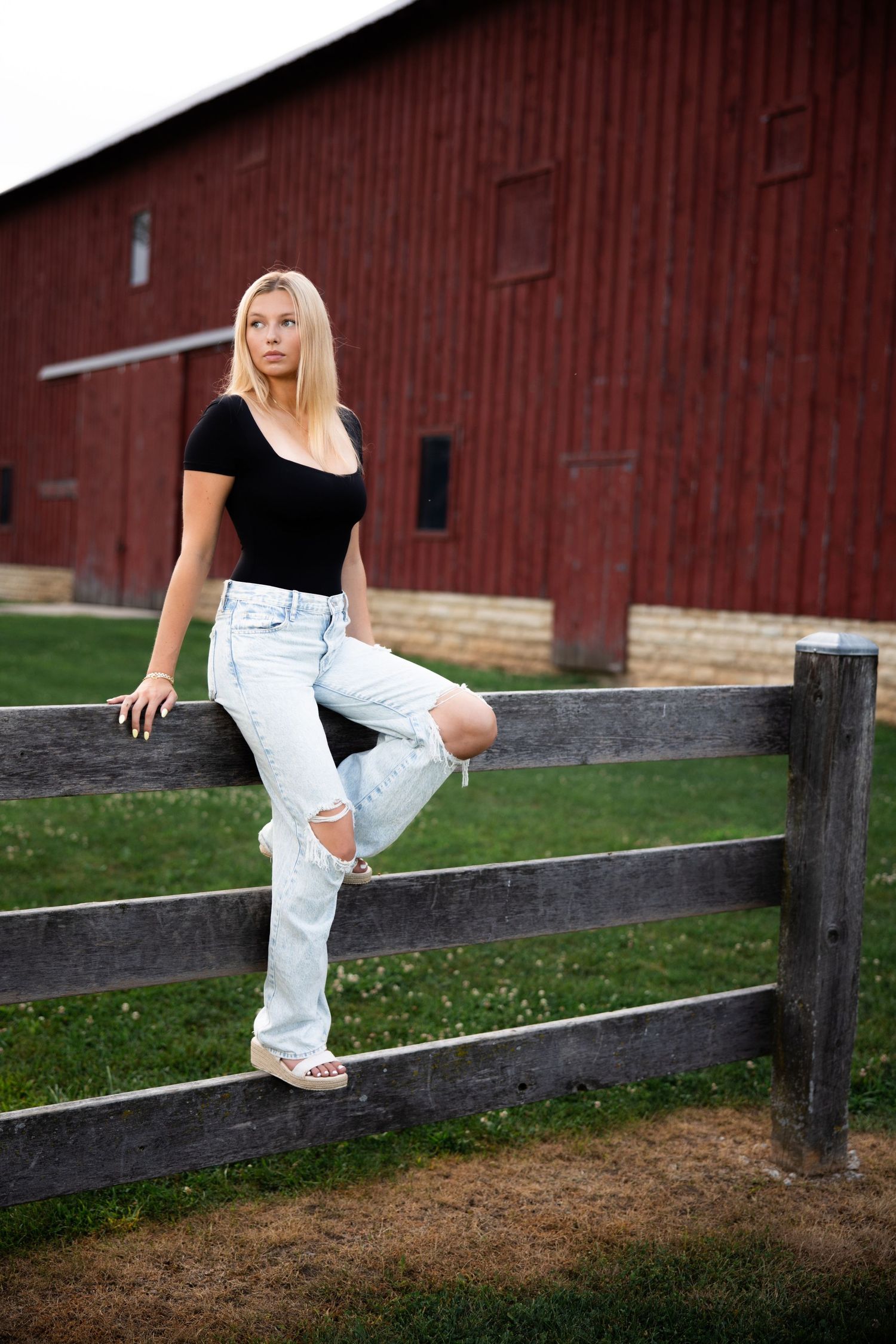 Figure in casual attire sits on a wooden fence with a rustic red barn in the background.