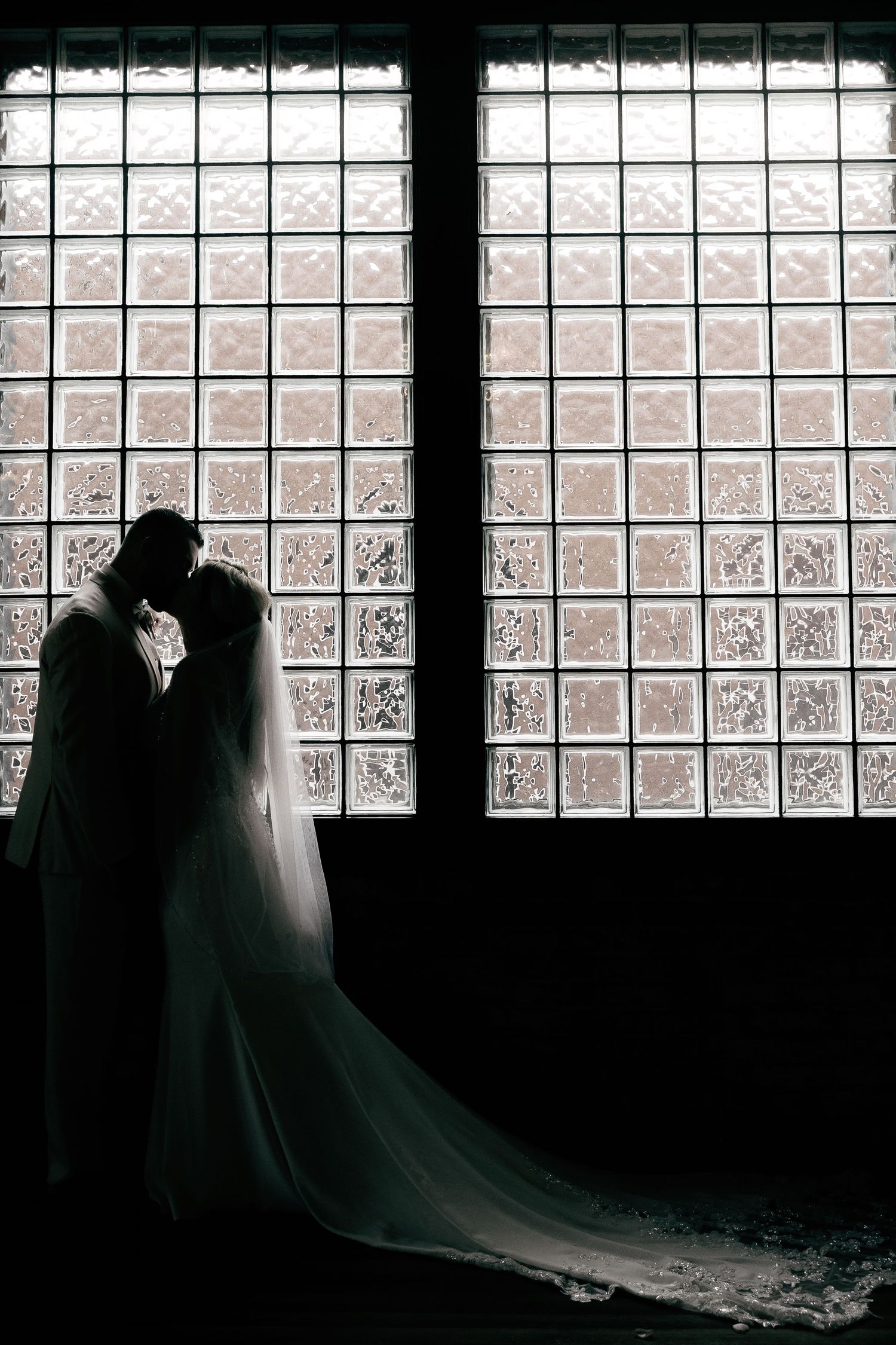 Silhouetted wedding couple poses against a window with dramatic lighting and glass block backdrop.