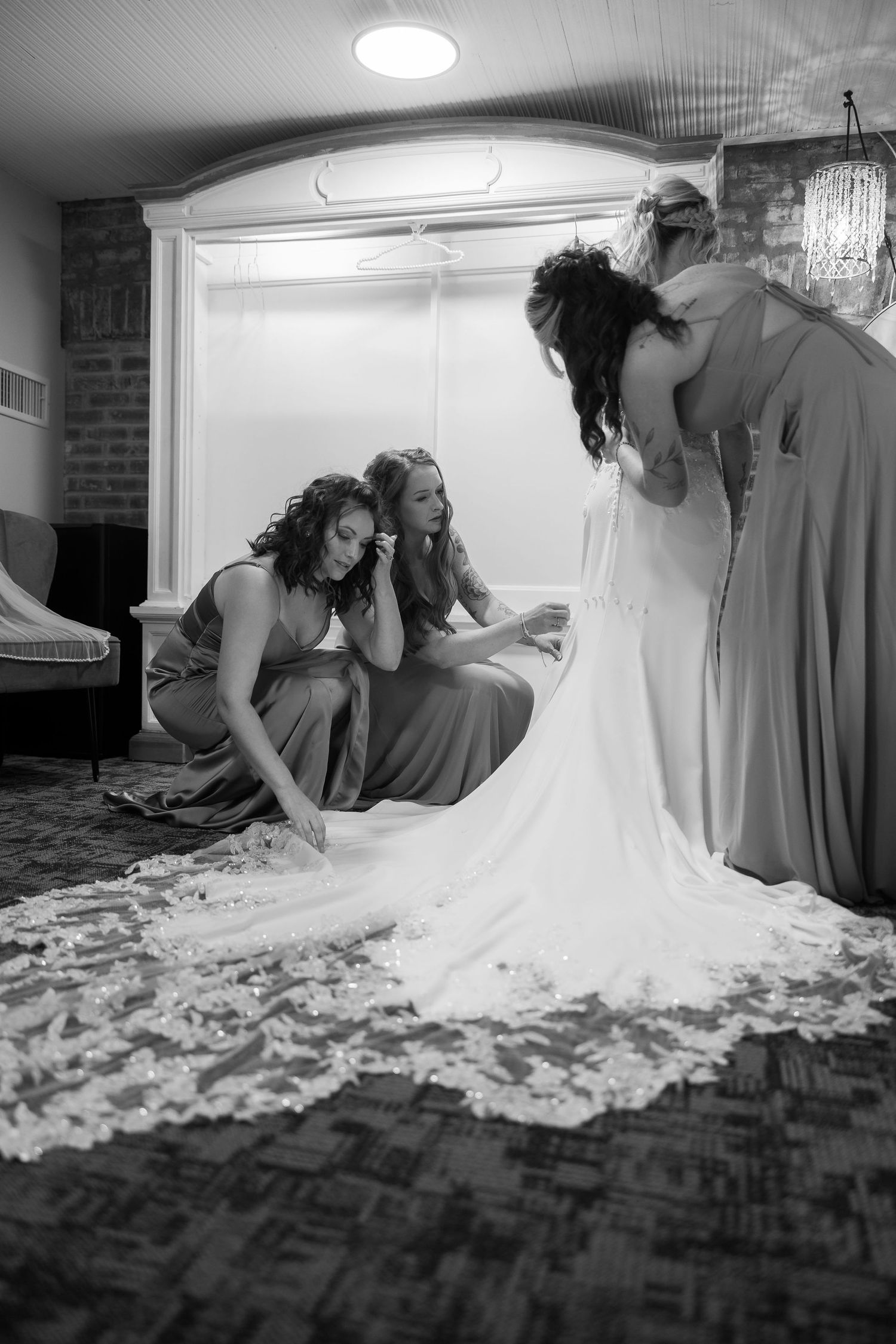 Black and white photo of bridesmaids arranging a wedding dress train on a hardwood floor.