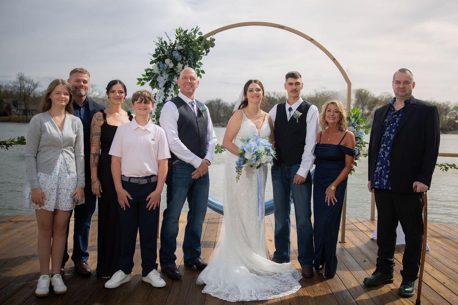 Wedding party poses together under a circular floral arch on a waterfront deck.