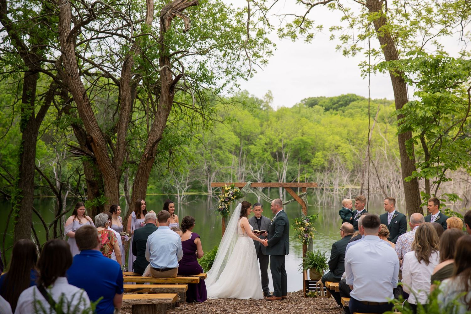 Outdoor wedding ceremony by a serene lake with guests seated on wooden benches among tall trees.