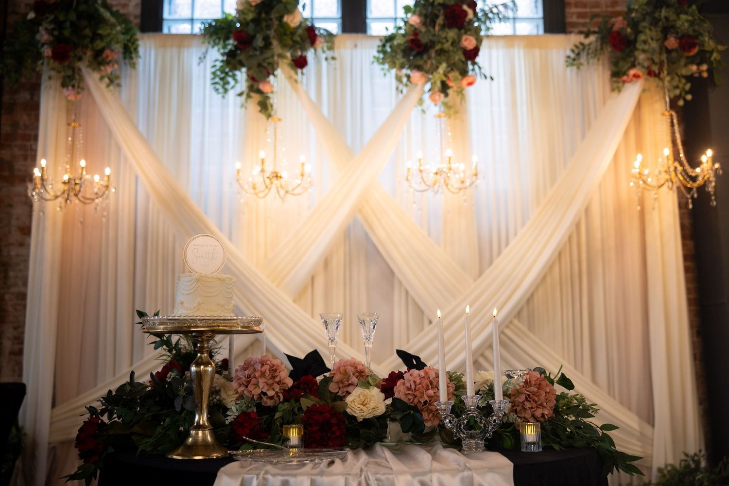 Elegant wedding reception backdrop with white draping, crystal chandeliers, floral arrangements, and a decorated sweetheart table.