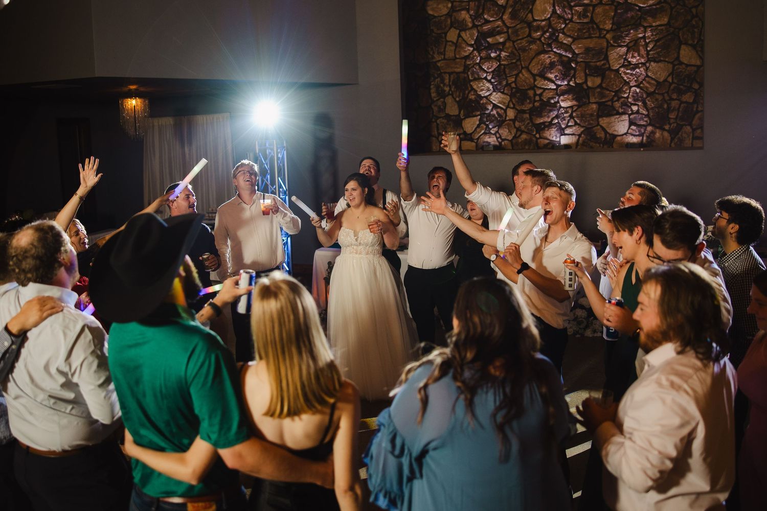 Wedding reception dance floor with bride and guests celebrating, holding glow sticks under dramatic lighting.