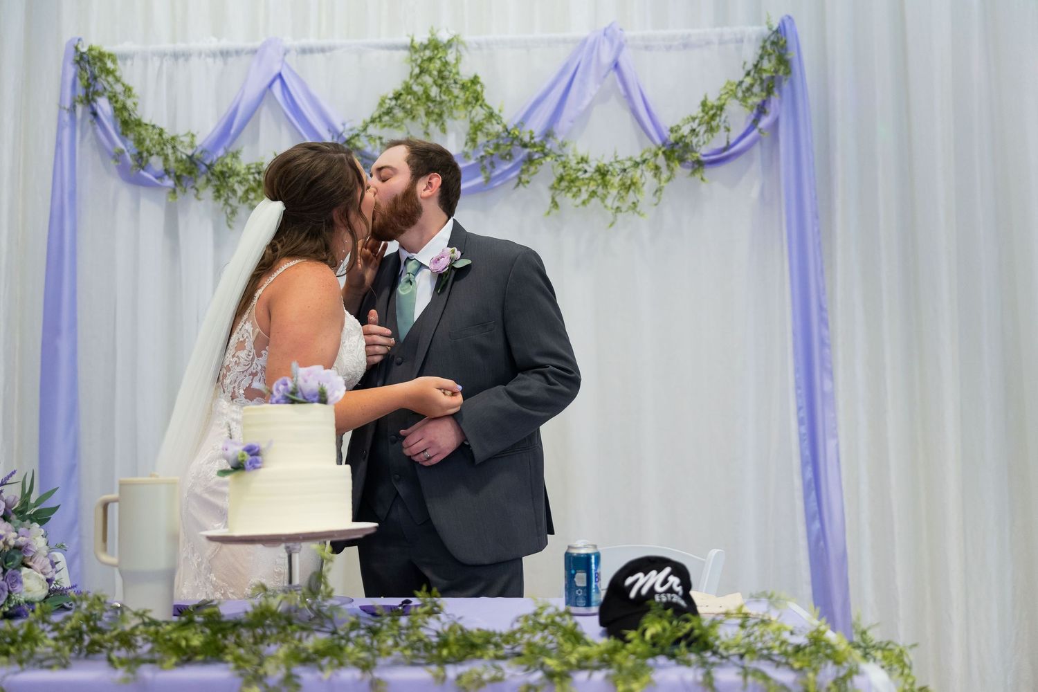 Bride and groom share a kiss while cutting their wedding cake at a reception with purple floral decorations.